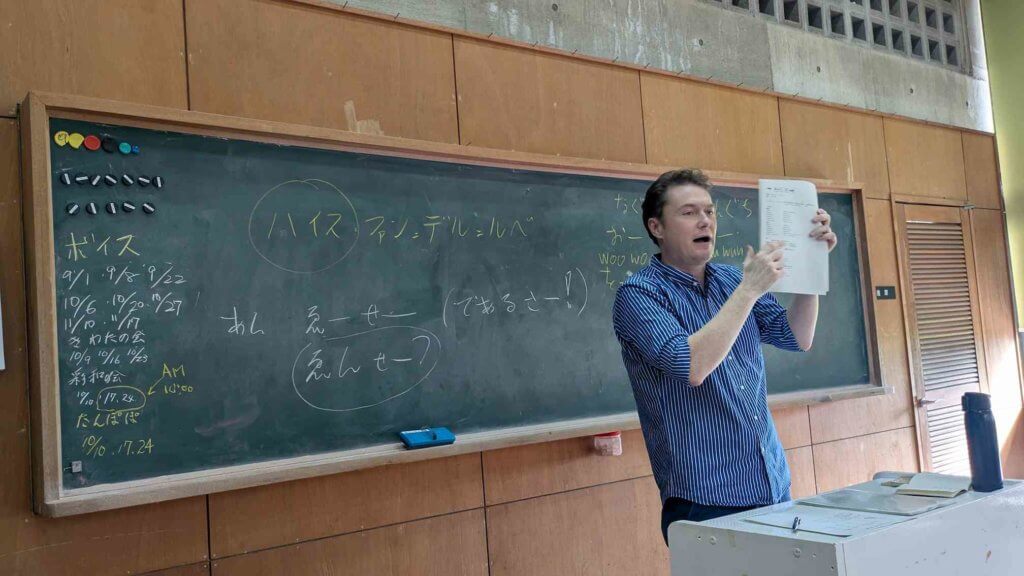 沖縄語講座のようす。講師のハイス氏が黒板の前に立ち、プリントを手にもってかざしている。